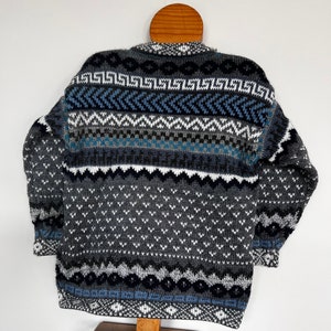 K&ouml;nnte beinhalten: Ein grauer, blauer und wei&szlig;er Pullover mit einem Rundhalsausschnitt. Der Pullover hat ein geometrisches Muster mit verschiedenen Formen und Farben.