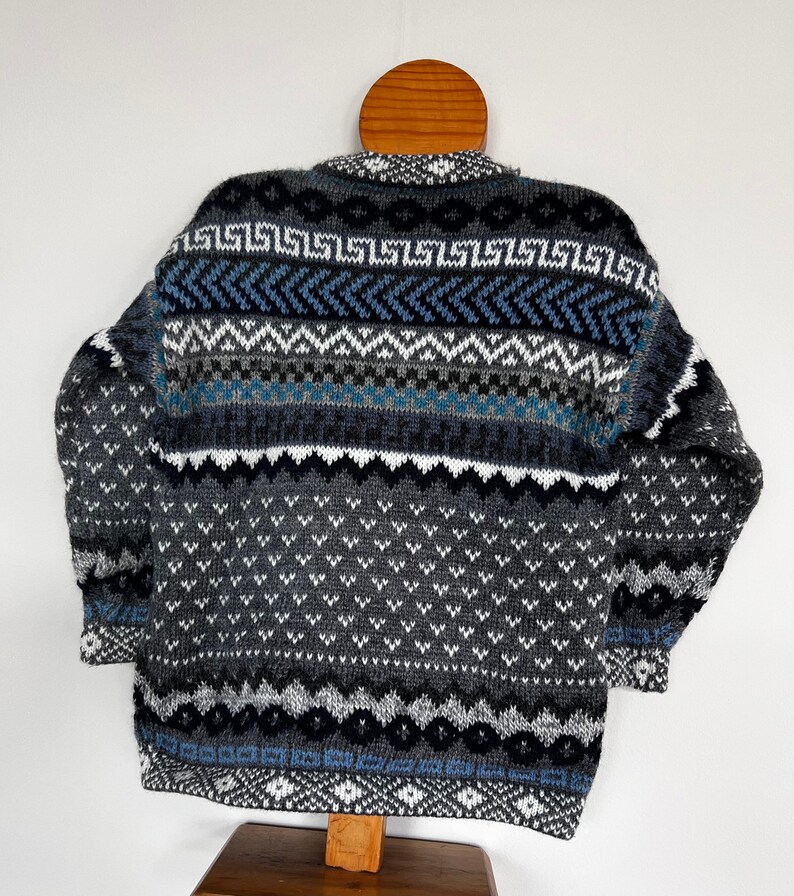 K&ouml;nnte beinhalten: Ein grauer, blauer und wei&szlig;er Pullover mit einem Rundhalsausschnitt. Der Pullover hat ein geometrisches Muster mit verschiedenen Formen und Farben.