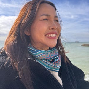 May include: A woman wearing a colorful knit cowl with a geometric pattern in teal, pink, and white. She is wearing a black jacket and smiling, with the ocean and sky in the background.