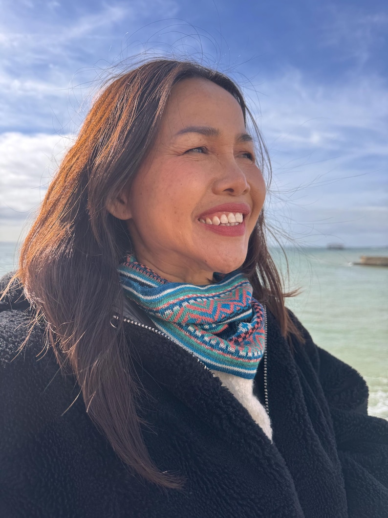 May include: A woman wearing a colorful knit cowl with a geometric pattern in teal, pink, and white. She is wearing a black jacket and smiling, with the ocean and sky in the background.