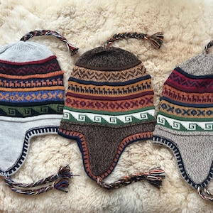 May include: Three knitted winter hats with ear flaps and colorful geometric patterns. The hats are made of wool and have a braided tassel on the top.