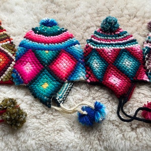 May include: Four colorful crocheted hats with pom-poms. The hats are made with a variety of colors, including green, blue, pink, red, and white. The hats have a diamond pattern and are all different sizes.