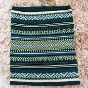 May include: A green and white patterned neck gaiter with a geometric design. The gaiter has a dark green top and bottom border, with a tan drawstring. The pattern includes diamonds, zigzags, and horizontal stripes.
