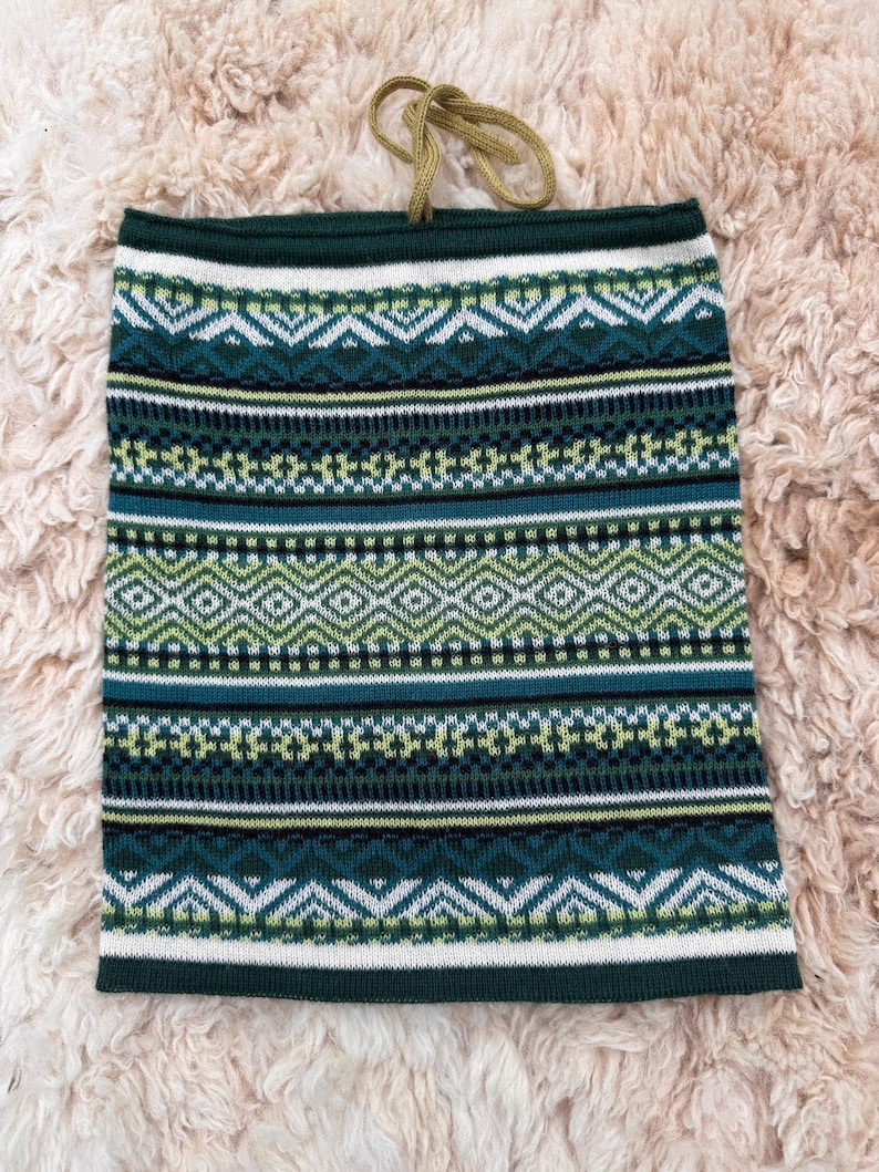 May include: A green and white patterned neck gaiter with a geometric design. The gaiter has a dark green top and bottom border, with a tan drawstring. The pattern includes diamonds, zigzags, and horizontal stripes.