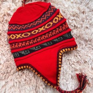 May include: A vibrant red knit hat with ear flaps and a braided tassel. The hat features intricate geometric patterns in yellow, orange, black, and purple. The hat is designed with a warm, cozy style.