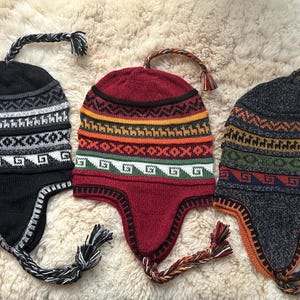 May include: Three colorful knit hats with ear flaps and braided ties. The hats feature geometric patterns in black, white, red, orange, green, and blue. The hats are displayed on a textured, cream-colored surface.