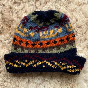 May include: A knitted beanie featuring a multi-coloured geometric pattern in shades of navy blue, orange, yellow, olive green, and burgundy. The hat has a wide, folded cuff at the bottom and a rounded crown, showcasing a textured, wool-like yarn construction.