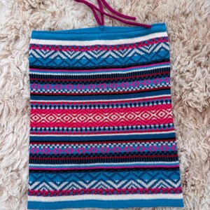 May include: A knitted neck gaiter with a vibrant geometric pattern in shades of blue, pink, white, and black. The neck warmer has a drawstring at the top and is designed to be worn around the neck. The fabric appears soft and warm.