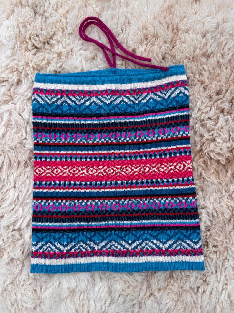 May include: A knitted neck gaiter with a vibrant geometric pattern in shades of blue, pink, white, and black. The neck warmer has a drawstring at the top and is designed to be worn around the neck. The fabric appears soft and warm.