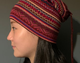 May include: A red, pink, brown, and blue patterned knit beanie with a long tassel. The beanie is worn by a person with long dark hair.