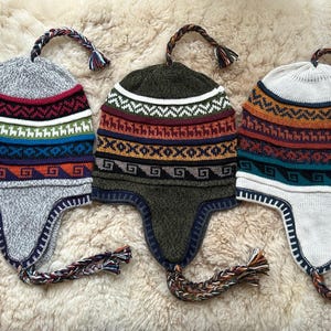 Op de afbeelding: Drie gebreide wintermutsen met oorflappen en kleurrijke geometrische patronen. De mutsen zijn grijs, groen en wit met een verscheidenheid aan kleuren in de patronen.