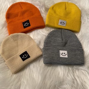 Può includere: Quattro berretti colorati in arancione, giallo, beige e grigio. Ogni berretto presenta una piccola toppa quadrata nera con una faccina sorridente bianca ricamata. I berretti sono esposti su una superficie bianca e soffice.