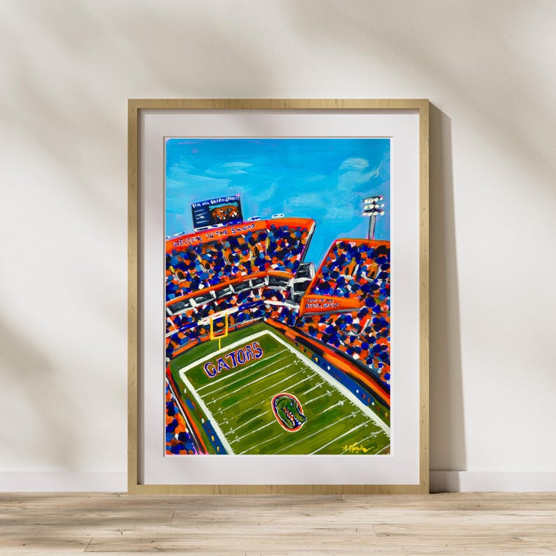 Pu&ograve; includere: Un dipinto colorato di uno stadio di calcio con un campo verde e la parola "GATORS" al centro. Le tribune sono piene di tifosi in arancione e blu. Il dipinto &egrave; incorniciato in una cornice di legno chiaro.