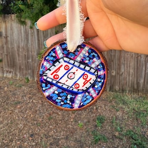 May include: A wooden ornament painted with a hockey rink and a crowd of fans. The ornament is painted in shades of blue, pink, white, and red.