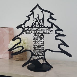 May include: A black metal sculpture of a cedar tree, the national symbol of Lebanon, with Arabic calligraphy inscribed on the trunk. The sculpture is on a black base.