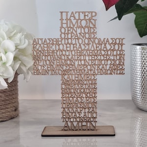 Pode incluir: Uma cruz de madeira com a ora&ccedil;&atilde;o do Senhor em grego escrita nela. As palavras s&atilde;o cortadas na madeira, criando uma pe&ccedil;a &uacute;nica e decorativa.