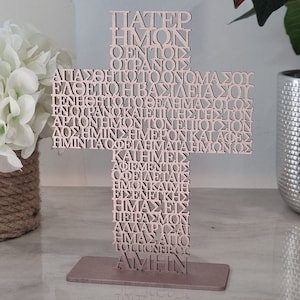 Pode incluir: Uma cruz de madeira com a ora&ccedil;&atilde;o do Senhor em texto grego, escrita em uma fonte decorativa. A cruz &eacute; pintada de uma cor rosa claro e fica em uma base de metal.