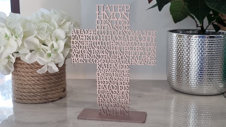 Pode incluir: Uma cruz de madeira com a ora&ccedil;&atilde;o do Senhor em texto grego, escrita em uma fonte decorativa. A cruz &eacute; pintada de uma cor rosa claro e fica em uma base de metal.