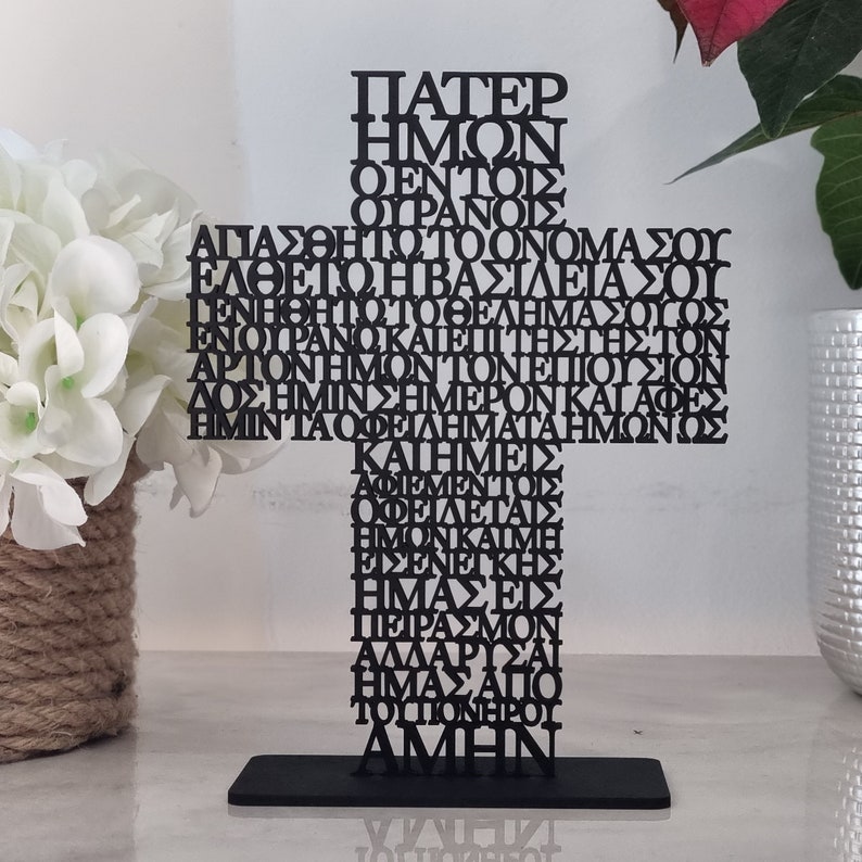 Pode incluir: Uma cruz de metal preto com a ora&ccedil;&atilde;o do Senhor em grego escrita nela. A cruz est&aacute; em p&eacute; sobre uma base preta.