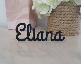 May include: Black wooden sign with the name "Eliana" written in a cursive font.