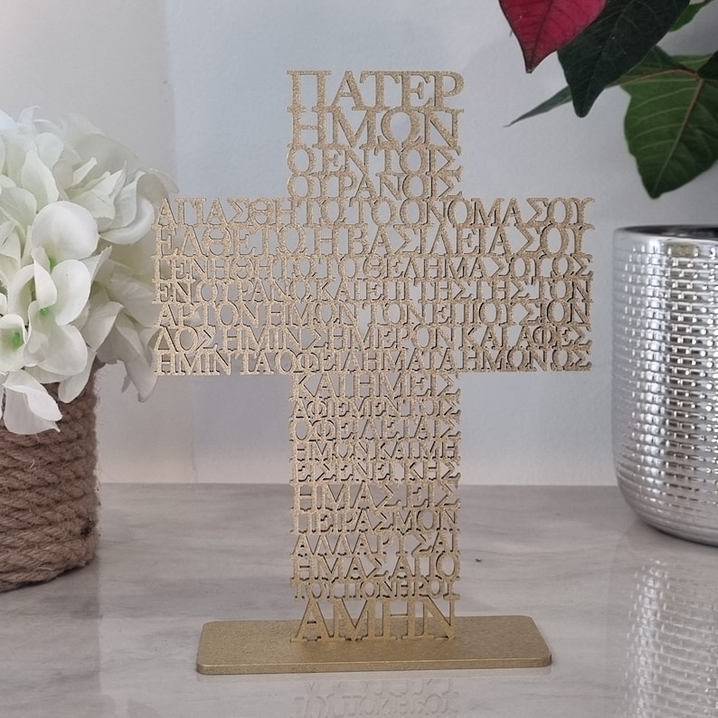 Pode incluir: Uma cruz de metal dourada com a ora&ccedil;&atilde;o do Pai Nosso em texto grego. A cruz est&aacute; sobre um suporte de metal dourado.