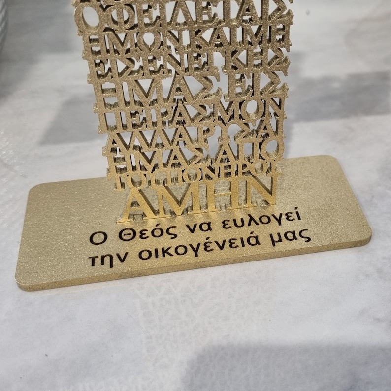 Pode incluir: Um suporte de metal dourado com um design de corte decorativo que apresenta as palavras gregas "&Omicron; &Theta;&epsilon;ό&sigmaf; &nu;&alpha; &epsilon;&upsilon;&lambda;&omicron;&gamma;&epsilon;ί &tau;&eta;&nu; &omicron;&iota;&kappa;&omicron;&gamma;έ&nu;&epsilon;&iota;ά &mu;&alpha;&sigmaf;", que se traduz como "Que Deus aben&ccedil;oe nossa fam&iacute;lia".