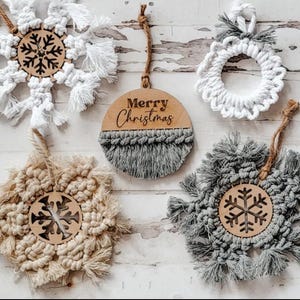 May include: A collection of handmade Christmas ornaments. The ornaments include macrame snowflakes, a wreath, and a wooden disc with "Merry Christmas" text. The ornaments are in neutral colors with wooden accents and hanging cords.
