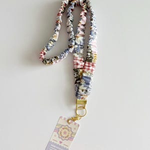 May include: A fabric lanyard featuring a gathered, scrunchie-style design with a patchwork pattern of floral, gingham, and solid pastel fabrics. It includes a gold-toned metal clasp and a rectangular tag with the text Marisa Creations Made With Love.