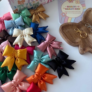 May include: A collection of colorful satin bows in various shades, including pink, blue, gold, and black, are scattered on a white surface. A wooden tray and a box with bow patterns are also visible. The bows are likely hair accessories or decorative elements.