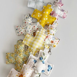 Puede incluir: Una colección de llaveros de tela con forma de cruz que presentan varios patrones, incluyendo cuadros vichy amarillos, estampados florales, un motivo de sol e ilustraciones de arándanos. Cada cruz acolchada incluye un mosquetón de metal plateado.