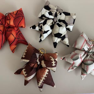 Op de afbeelding: Een verzameling stoffen strikvormige sleutelhangers met sportbalpatronen. Ontwerpen omvatten basketbal, voetbal, American football en honkbal. Elke strik heeft een metalen sleutelhanger.