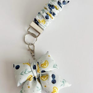 May include: A white fabric keychain with a bow and wristlet. The bow and wristlet are decorated with a pattern of yellow lemons, blue berries, and green leaves. The keychain has a silver metal clasp and key ring.