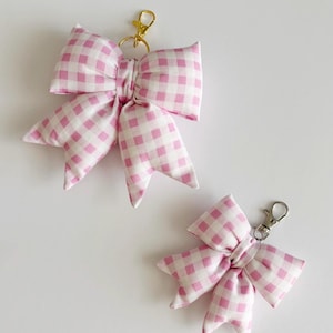 May include: Two fabric bows with a pink and white gingham pattern, each featuring a metal lobster clasp keychain attachment. The larger bow has a gold-toned metal clasp, while the smaller bow features a silver-toned metal lobster clasp.