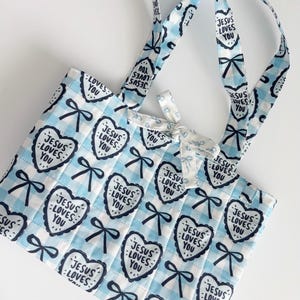 May include: A quilted tote bag featuring a light blue and white gingham pattern. The fabric is decorated with a repeating print of dark blue bows and heart-shaped outlines containing the text Jesus Loves You. It includes two handles and a bow.