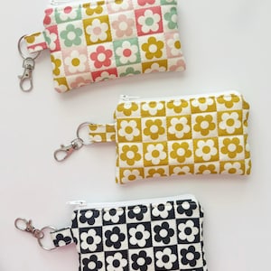 May include: Three rectangular zippered pouches with a quilted checkerboard pattern featuring flower motifs. The top pouch has a multi-colored floral grid, the middle is mustard yellow and white, and the bottom is black and white. Each has a metal keychain.