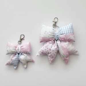 May include: Two fabric bow keychains in different sizes, featuring a patchwork design of light pink, white, and blue floral and gingham patterns. Each bow is attached to a silver-toned metal lobster clasp and a circular split ring for attachment.