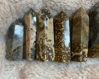 May include: Seven polished, pointed, natural stone crystals in various shades of brown, gray, and white. The crystals are arranged in a row on a white, fuzzy surface.