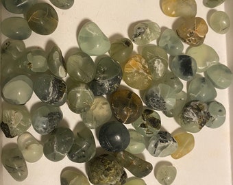 May include: A collection of green, gray, and brown tumbled stones with black and white inclusions. The stones are smooth and polished, and they are arranged in a random pattern.