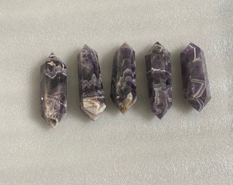 May include: Five amethyst crystal points with a purple and white striped pattern. The points are arranged in a row on a white surface.