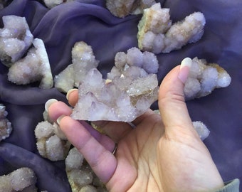 May include: A collection of small, white, amethyst crystal clusters with a purple hue. The crystals are arranged on a purple fabric background.