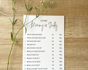 Peut inclure: Une carte blanche avec le texte "Guess Who? Mommy or Daddy" en écriture élégante, avec une liste de questions pour déterminer qui fera quoi. De délicates fleurs blanches et de la verdure sont disposées sur une surface en bois clair.