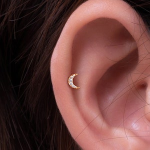 May include: A close-up of a person's ear with a gold crescent moon earring with small clear stones.