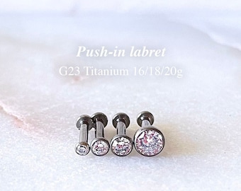 Pode incluir: Cinco pinos de labrete de titânio com topos de cristal transparente. Os pinos são de diferentes tamanhos, calibre 16, calibre 18 e calibre 20. O texto na imagem diz "Push-in labret G23 Titanium 16/18/20g".