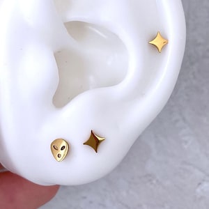 May include: A pair of gold earrings, one featuring a small alien face design and the other a small diamond shape.