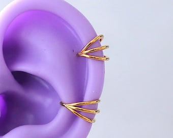 Pode incluir: Um close-up de um modelo de orelha roxo com três brincos de argola dourados. Os brincos de argola estão empilhados um em cima do outro e são feitos de fio fino.