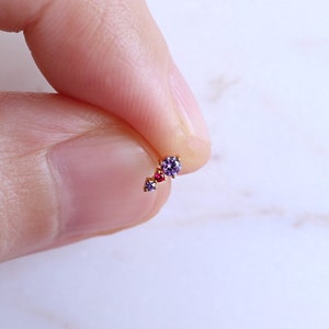 May include: A small gold bar with three tiny gemstones. The gemstones are purple and red.