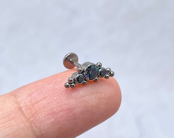 May include: A silver body piercing with a black gem and smaller black gems on either side. The piercing is on a finger.