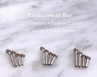 Pode incluir: Um conjunto de cinco barras de labret de titânio prateado de diferentes comprimentos. As barras estão dispostas em uma fila, com a barra mais curta à esquerda e a barra mais longa à direita. O texto "Replacement Bar" está escrito acima das barras, e o texto "G23 Titanium Push-in Labret 16/18/20g 5/6/8/10mm" está escrito abaixo das barras.