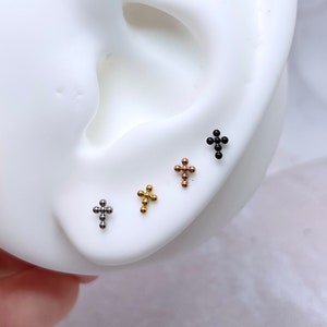 May include: Four small cross-shaped earrings in different colors: silver, gold, rose gold, and black. The earrings are shown on a white ear model.