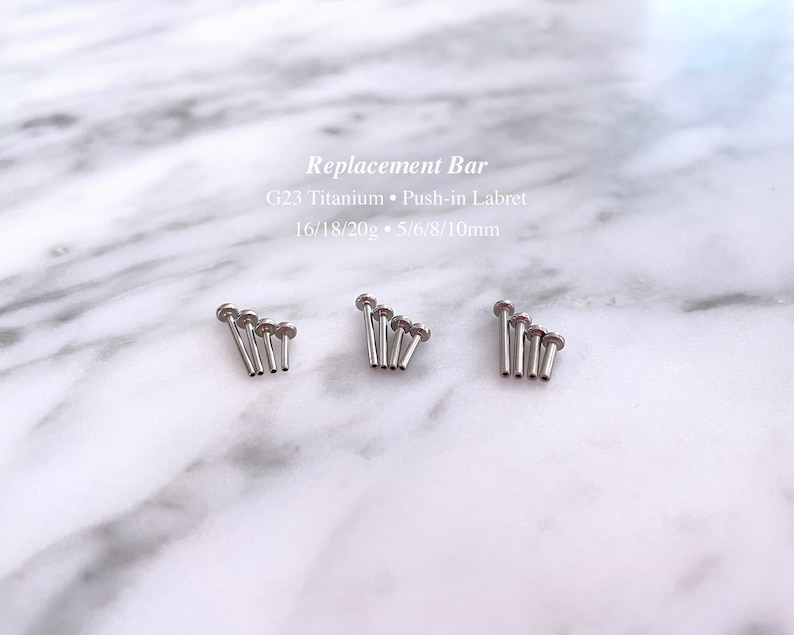 May include: A set of five silver titanium push-in labret bars in different lengths. The bars are arranged in a row, with the shortest bar on the left and the longest bar on the right. The text "Replacement Bar" is written above the bars, and the text "G23 Titanium Push-in Labret 16/18/20g 5/6/8/10mm" is written below the bars.